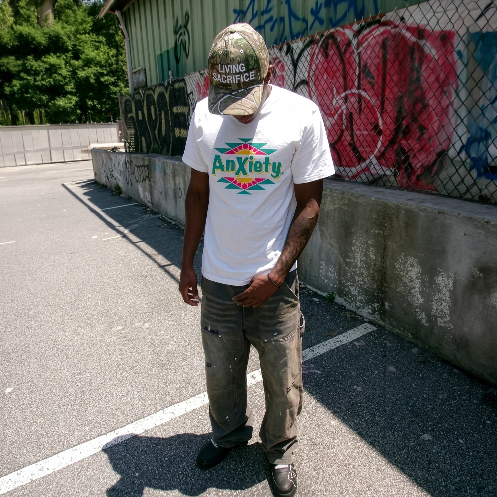 A man standing outdoors in a concrete lot wearing a white graphic palm treat t-shirt with a colorful 'Anxiety' design, gray pants, and a camouflage baseball cap that reads 'Living Sacrifice'.