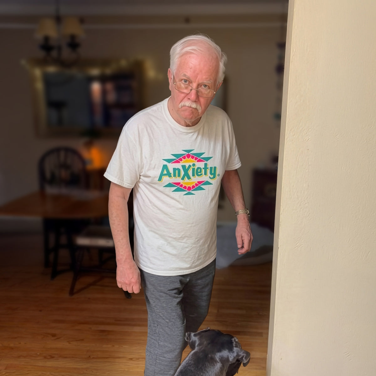 An older boomer man wearing a Palm Treat white graphic t-shirt with the word 'Anxiety' in colorful letters walks indoors with a black dog. The background shows a home interior with a wooden floor, dining table, and chairs.