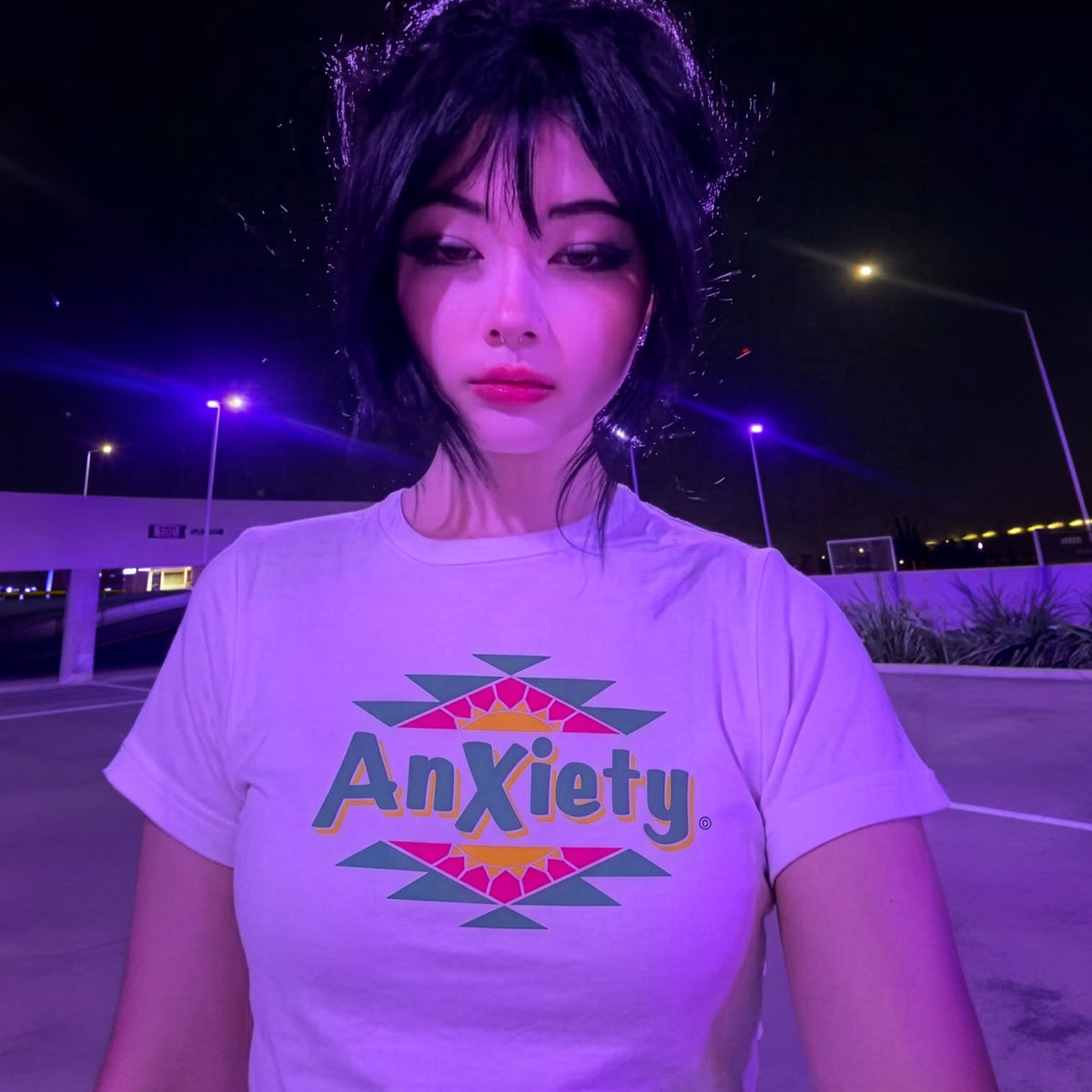A person wearing a white graphic palm treat T-shirt with the word 'Anxiety' in colorful letters and geometric shapes, photographed outdoors at night under purple streetlights.