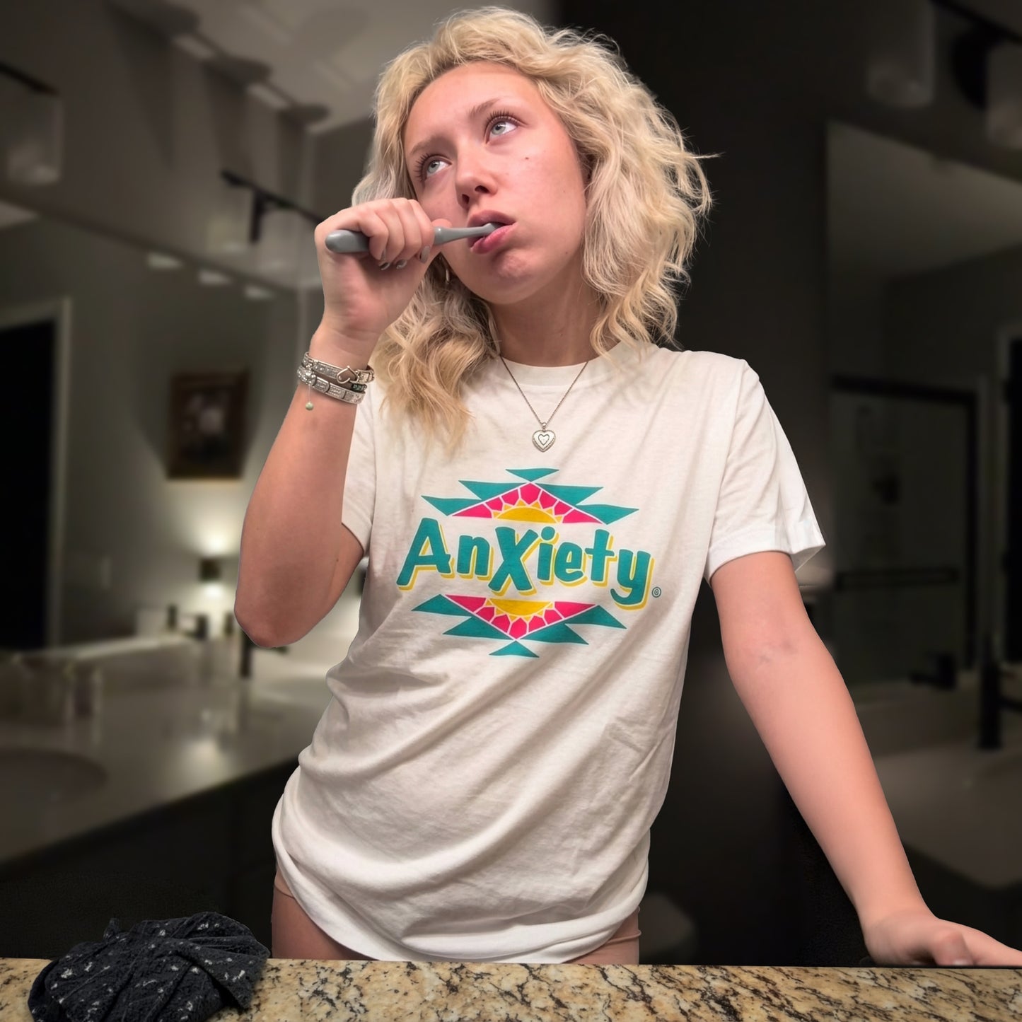 A woman with blonde hair brushing her teeth in a bathroom while wearing a white graphic palm treat t-shirt that reads 'Anxiety' in green with pink and teal geometric accents.