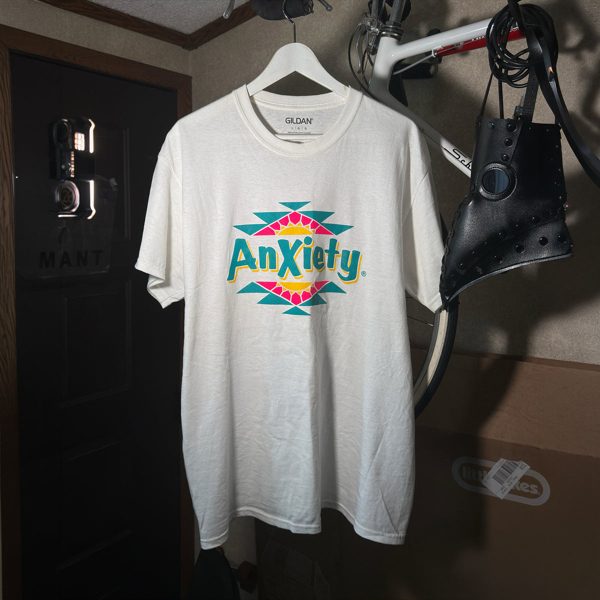 A white palm treat t-shirt hanging on a white hanger in a home setting, featuring a colorful graphic on the chest that reads 'Anxiety' inside a pink, yellow, and teal geometric design.